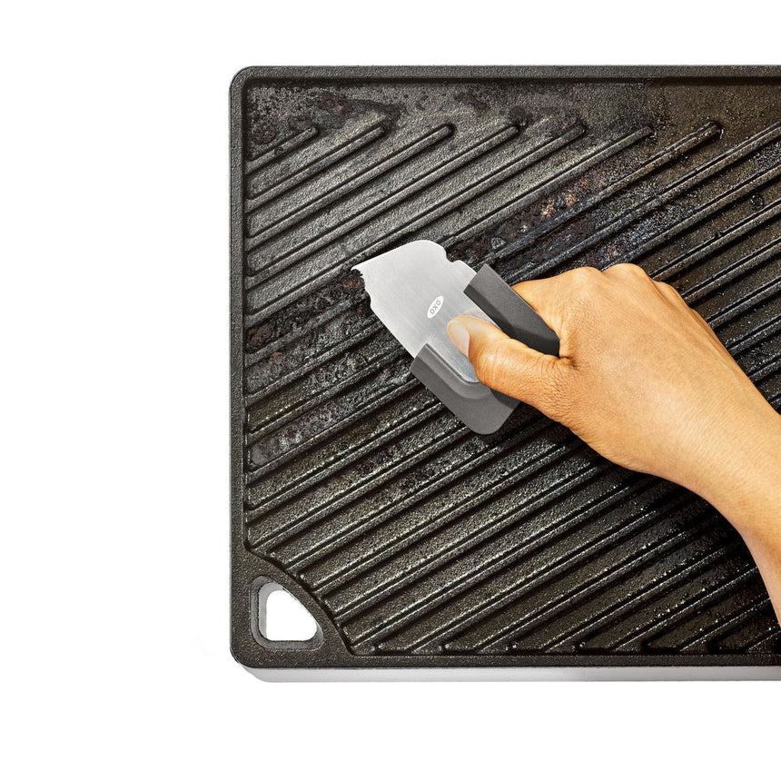Cast iron grill pan with ridges being cleaned by a hand using a scraper tool.