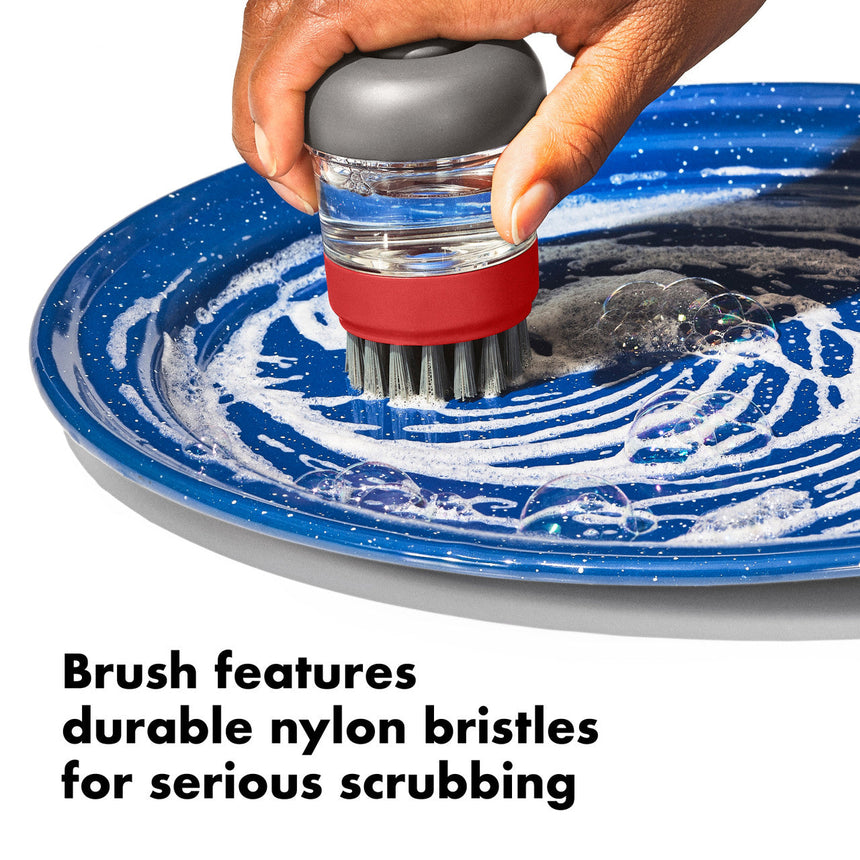 Handheld dish scrubbing brush with gray top, clear middle, and red base being used on a blue plate.