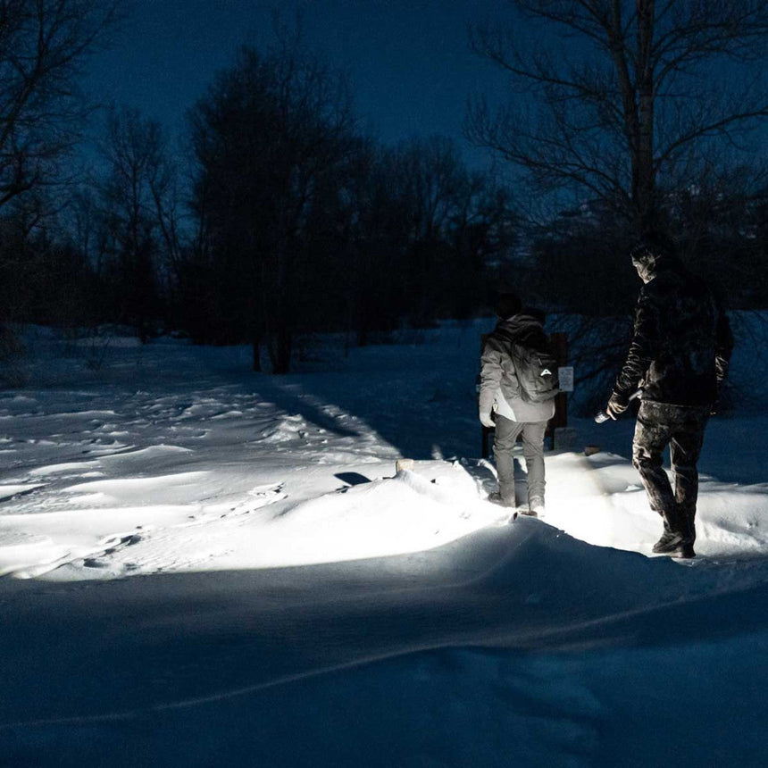 Two people walking through deep snow at night, illuminated by a bright light.