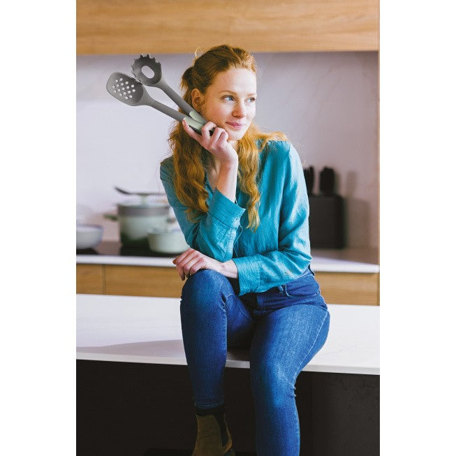 Woman with long braided hair wearing a teal sweater and jeans, holding a kitchen utensil.