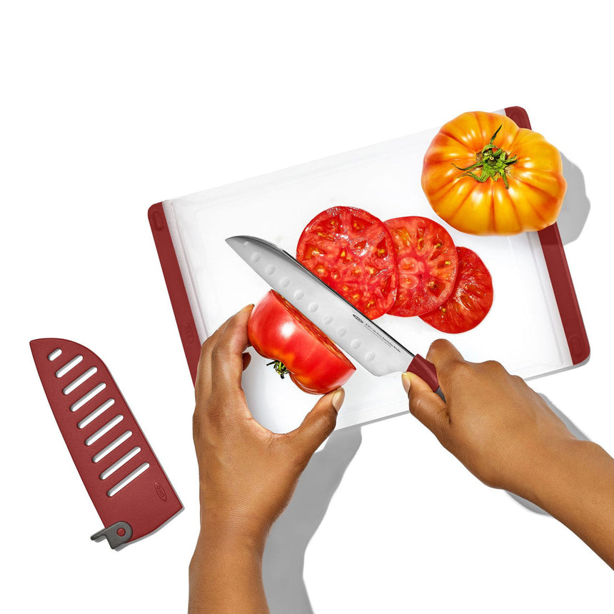 Hands slicing a tomato on a cutting board with a knife, alongside whole tomatoes.