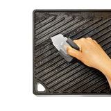 Cast iron grill pan with ridges being cleaned by a hand using a scraper tool.
