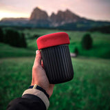 Reusable coffee cup with a red lid held up against a scenic mountain landscape.