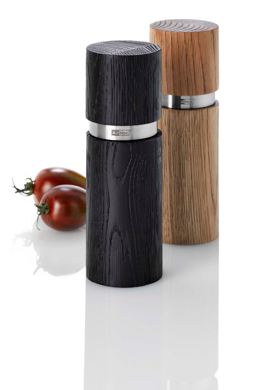 Wooden pepper mills with metal bands, one black and one light-colored, alongside two tomatoes.