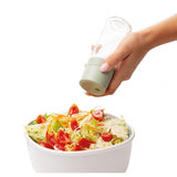 Salad bowl with mixed vegetables being seasoned from a shaker held by a hand.