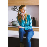Woman with long braided hair wearing a teal sweater and jeans, holding a kitchen utensil.